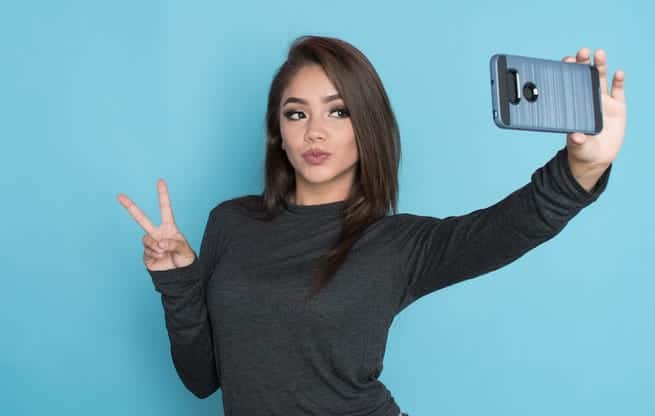 Brunette woman in front of blue background taking selfie to show off full, plump lips after fillers
