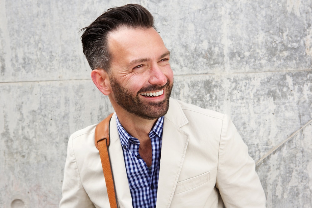 A portrait of a handsome smiling man in front of a concrete wall with styled hair and wearing a professional shirt and blazer