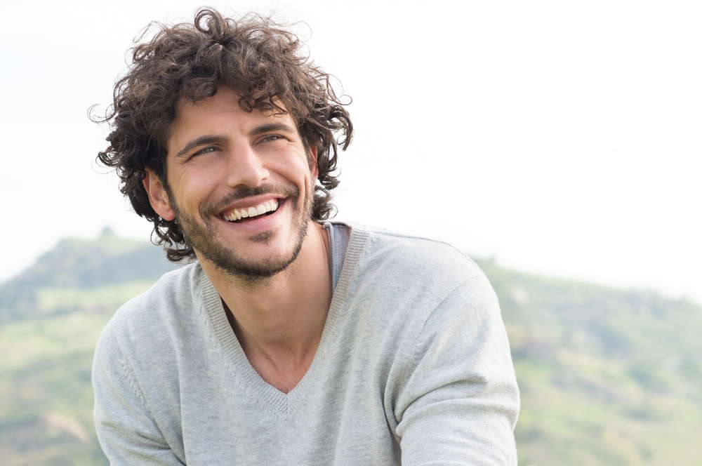 An attractive man with wavy full hair smiles widely while seated outside with a v-neck sweater on